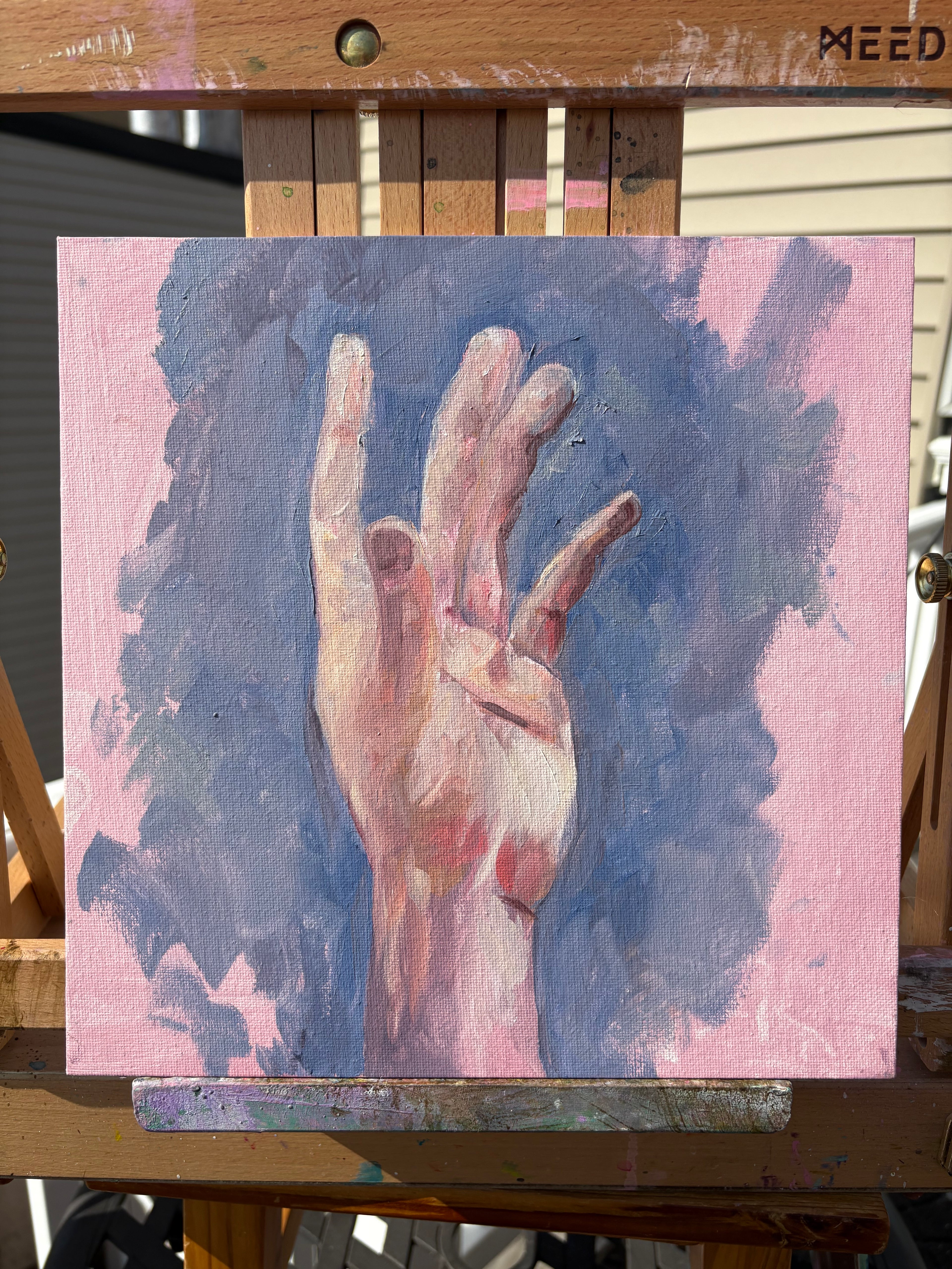 Oil painting on canvas panel by Natalie Blair - Brooklyn Artist, displayed on a wooden easel outdoors, depicting a hand reaching upward against a pink and blue background, in natural daylight showing the texture of the canvas and brushwork.