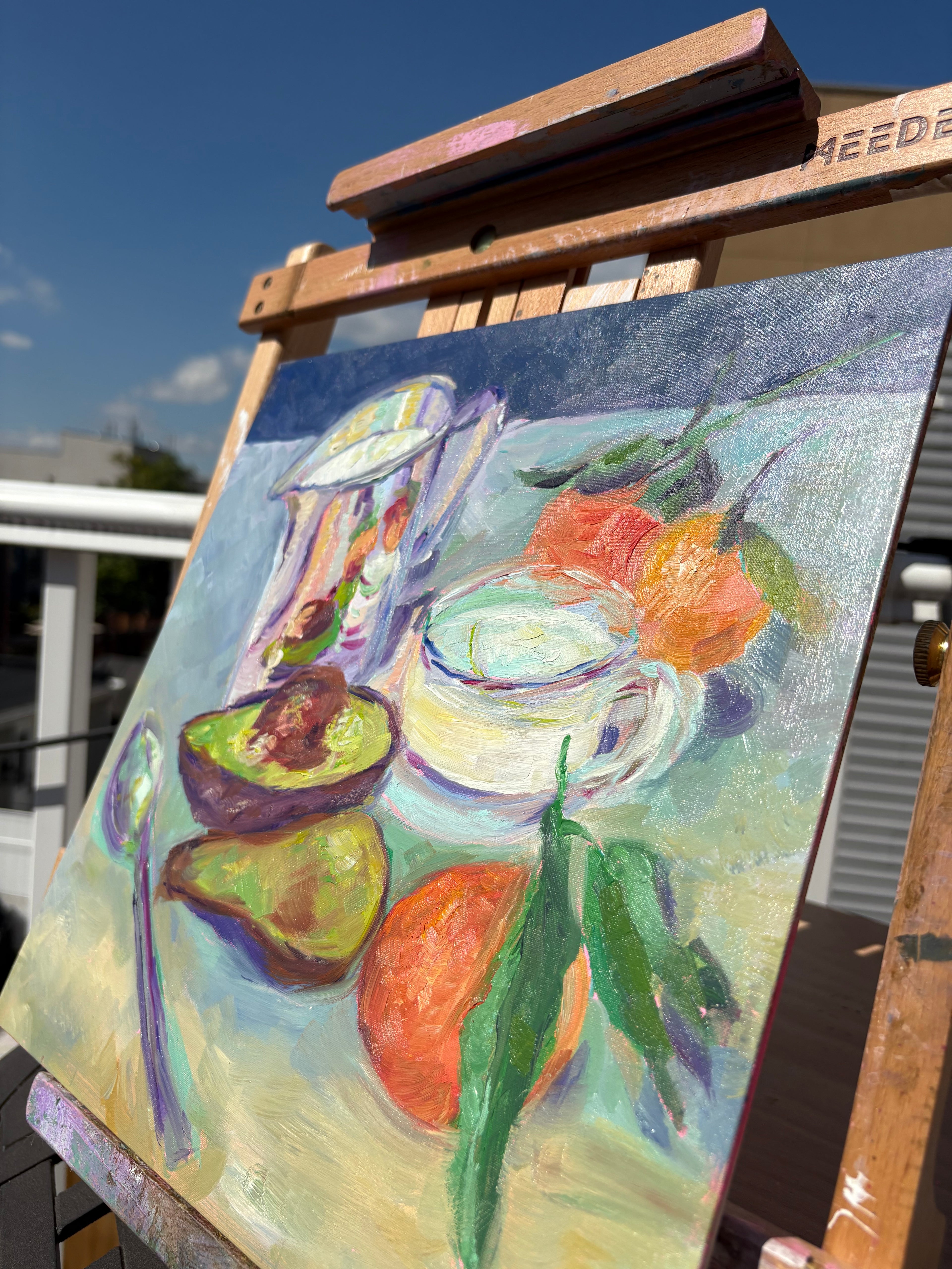 Angled detail image of an oil painting of an impressionist style still life with a shiny pitcher, halved avocado, cup of milk, and oranges.