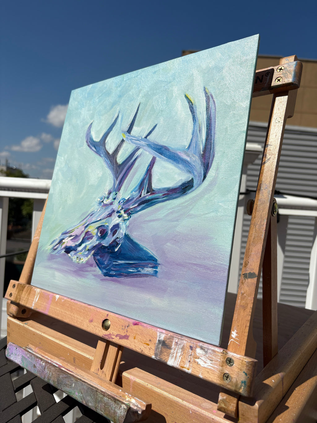Colorful oil painting of a deer skull with a large rack of antlers, leaning on a book - colors are jewel-toned pinks, purples, yellows and blues