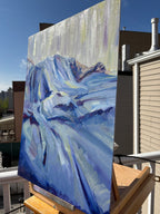 Oil painting on canvas panel displayed on a wooden easel on a rooftop deck, showing the thick impasto texture of blue and white bedclothes in raking light, with Brooklyn rooftops and a clear blue sky behind it.