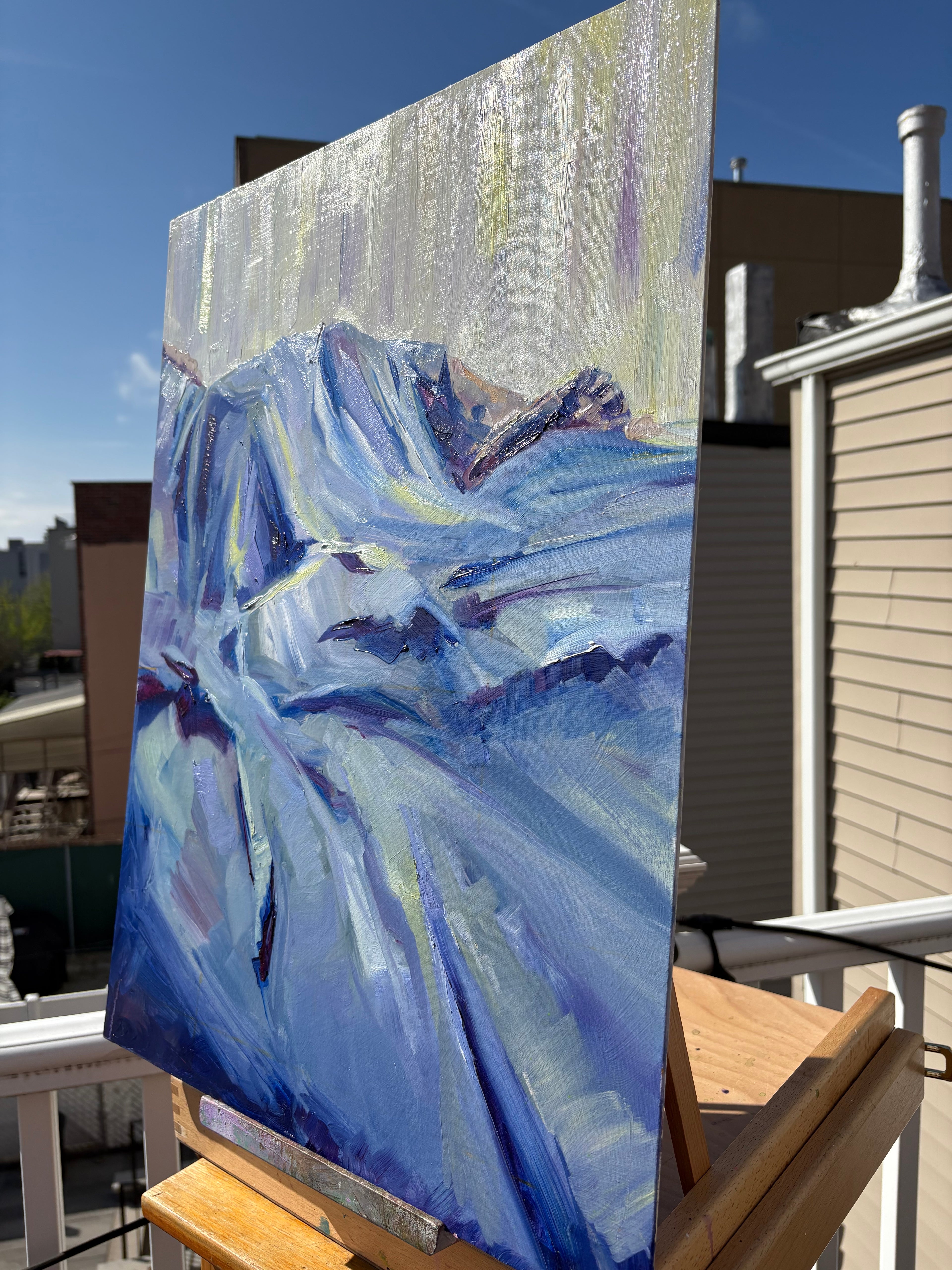 Oil painting on canvas panel displayed on a wooden easel on a rooftop deck, showing the thick impasto texture of blue and white bedclothes in raking light, with Brooklyn rooftops and a clear blue sky behind it.